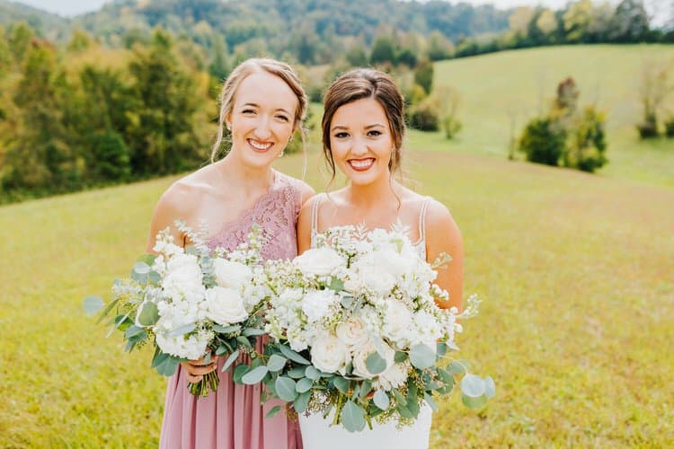 Bride and bridesmaid with white bouquets in a grassy field with trees, reflecting a Smoky Mountains outdoor wedding.