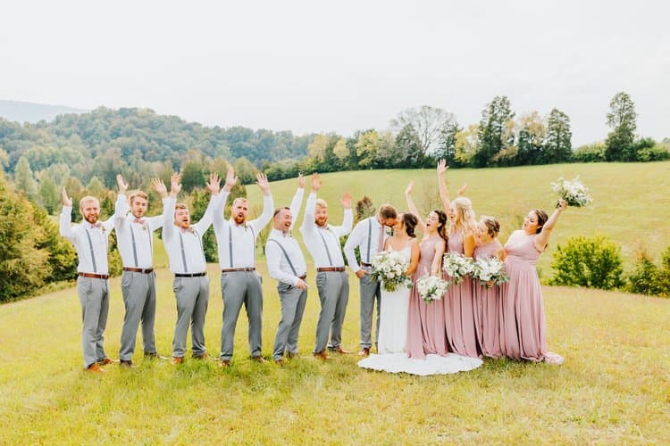 Wedding party with bride, groom, bridesmaids in mauve dresses, and groomsmen in suspenders in a grassy field with mountains.