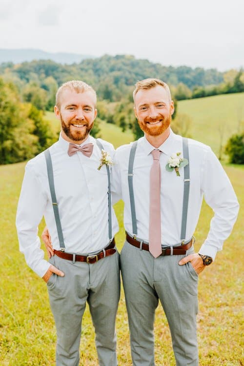 Two men in white shirts, grey suspenders, and pastel ties stand smiling in a grassy field with forested hills.