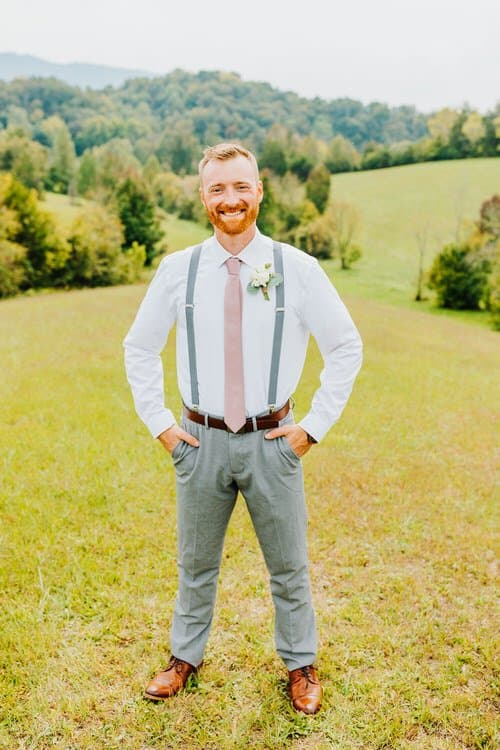 Groom in white shirt, grey suspenders, pink tie, and boutonniere stands in a grassy field with rolling hills and trees.